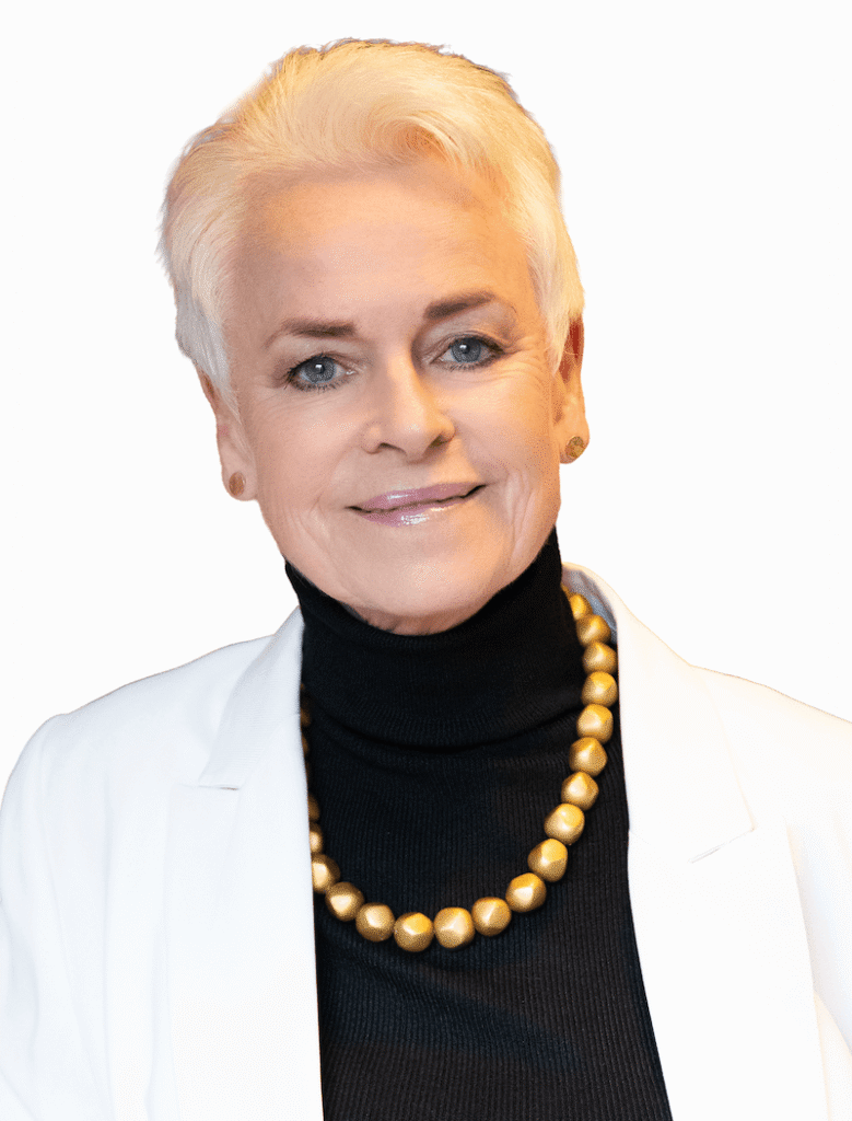 Portrait of a mature woman with short white hair, wearing a black turtleneck, white blazer, and gold beaded necklace against a white background.