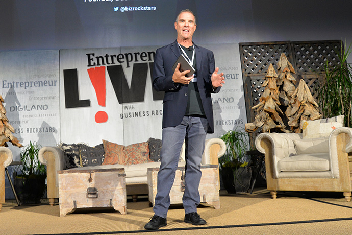 Male speaker on stage at Entrepreneur LIVE, gesturing with a tablet while presenting; backdrop shows ‘Entrepreneur LIVE’ and audience area with couches and decor behind him.